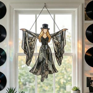 May include: A stained glass suncatcher depicting a figure wearing a top hat and a long, flowing black dress with wide sleeves. The figure has blonde hair and is suspended by a metal chain in front of a window. The glass features marbled textures.