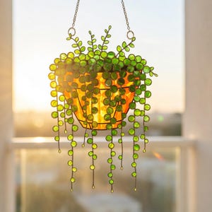 May include: Stained glass hanging planter with a string of pearls plant design. The planter is a warm yellow color, and the plant features green, round glass beads. The planter hangs from a chain, creating a decorative window accent.