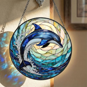 May include: Round stained glass panel featuring a blue dolphin leaping through a wave. The dolphin is detailed with shades of blue, and the water is a mix of blues and greens. The panel is framed in black and hangs from a chain.