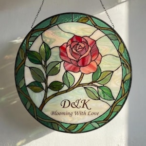 May include: Round stained glass panel featuring a red and pink rose with green leaves and vines. The panel has a green border and the text "D&K Blooming With Love". The panel is suspended by a chain.