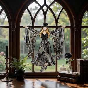 May include: Stained glass art piece depicting a woman in a black dress and top hat with outstretched arms. The artwork is in shades of gray and black, hanging in front of a window. A black rose in a vase and books are on the wooden surface.