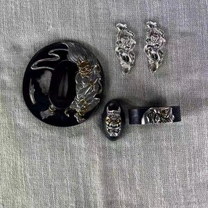 May include: A collection of Japanese sword fittings, including a black tsuba with a silver and gold demon design, two silver menuki, a black fuchi, and a kashira. The fittings are intricately detailed.
