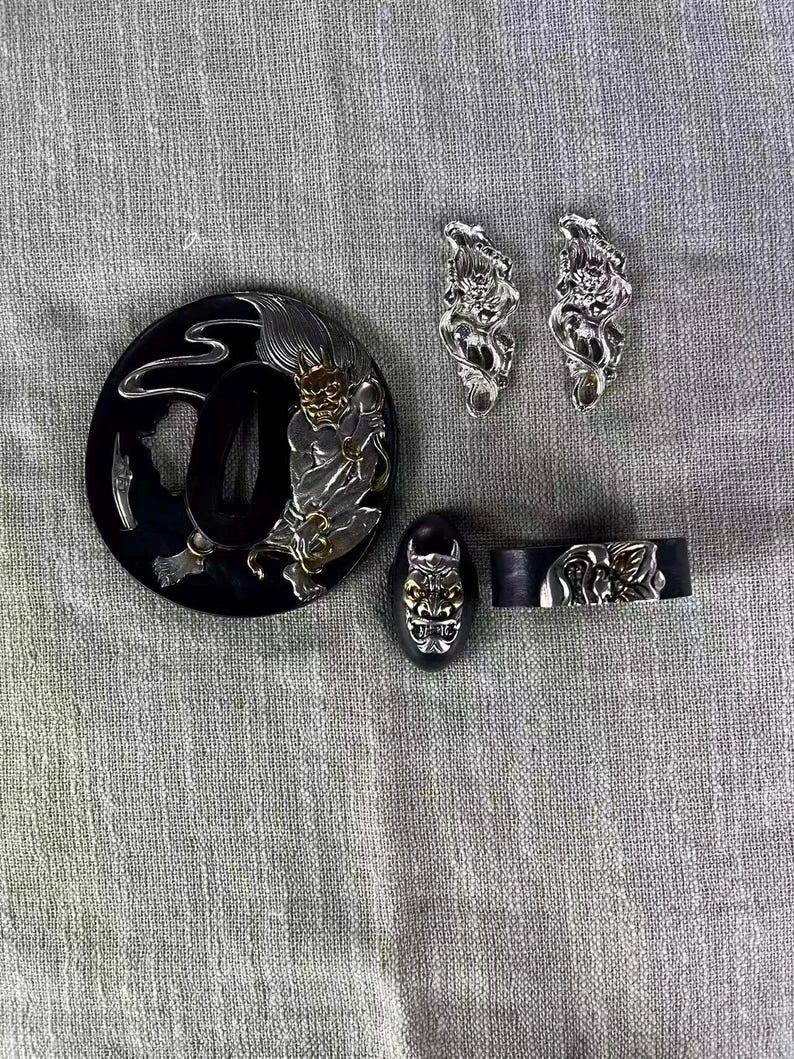 May include: A collection of Japanese sword fittings, including a black tsuba with a silver and gold demon design, two silver menuki, a black fuchi, and a kashira. The fittings are intricately detailed.