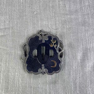May include: A decorative Japanese katana sword tsuba, featuring a dark blue central design with a gold crescent moon and bamboo accents. The tsuba has a silver-colored border and a central opening for the sword's tang.