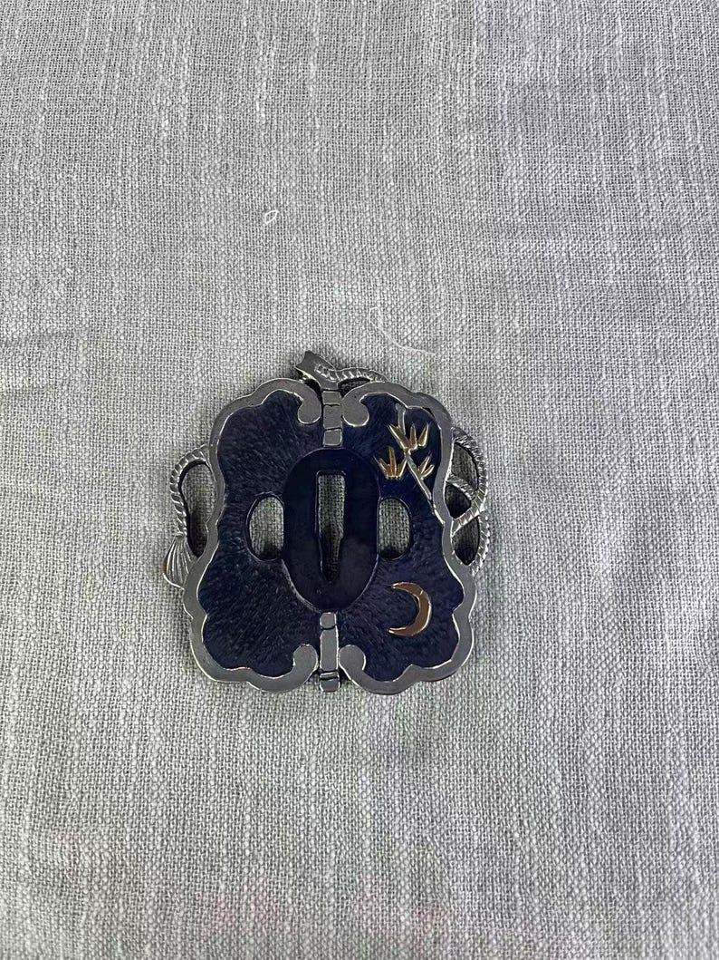 May include: A decorative Japanese katana sword tsuba, featuring a dark blue central design with a gold crescent moon and bamboo accents. The tsuba has a silver-colored border and a central opening for the sword's tang.