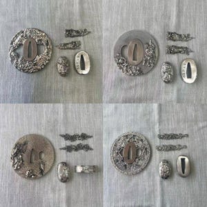 Op de afbeelding: Een verzameling antieke Japanse zwaardfittingen, waaronder tsuba, fuchi en kashira. De zilverkleurige metalen onderdelen tonen ingewikkelde bloemen- en geometrische patronen. De collectie wordt gepresenteerd op een neutraal gekleurd oppervlak.