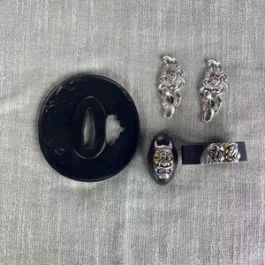 May include: A collection of Japanese sword fittings, including a black tsuba with a cutout, two silver menuki, a black fuchi with a gold mask design, and a black kashira with a silver and gold design.