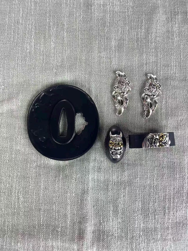 May include: A collection of Japanese sword fittings, including a black tsuba with a cutout, two silver menuki, a black fuchi with a gold mask design, and a black kashira with a silver and gold design.