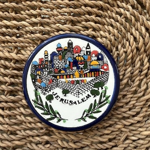 May include: A round, ceramic Jerusalem souvenir plate with a blue rim. The plate features a colorful illustration of the city, with the word "Jerusalem" in white text. Green olive branches are at the bottom.