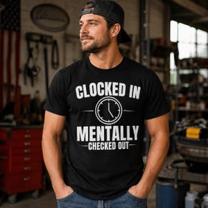 May include: Black t-shirt with the text "Clocked In Mentally Checked Out" in white. The design includes a clock graphic. The shirt is worn by a person wearing a black cap.