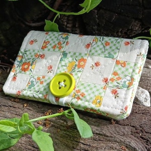 May include: A rectangular quilted fabric wallet featuring a patchwork pattern of white, light green gingham, and small orange and yellow floral prints. The wallet is secured with a single, bright lime green plastic button closure.