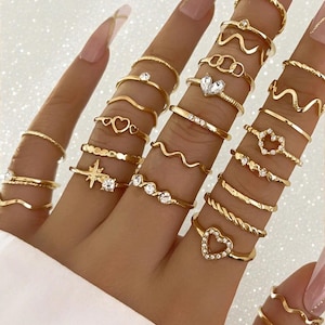 May include: A collection of gold-colored rings in various designs, including hearts, waves, and geometric shapes. Some rings feature clear gemstones. The rings are displayed on fingers, showcasing the different styles and textures of the jewelry.
