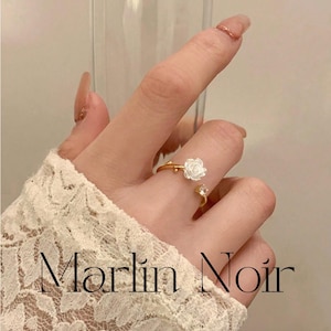 Peut inclure: Bague dorée ornée d'une rose blanche et d'une petite pierre transparente. La bague est réglable. Le texte "Marlin Noir" est visible sur l'image.