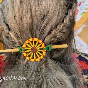 May include: A handmade hair accessory featuring a yellow and brown sunflower design. The hair pin is made of wood and green thread. The hair accessory is set against gray braided hair.