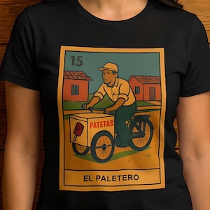 May include: Black t-shirt featuring a vintage-style illustration of a man on a bicycle selling paletas. The image includes the number 15, the word "PATETAS", and "EL PALETERO" in a rectangular frame. The design is in shades of yellow, green, and red.