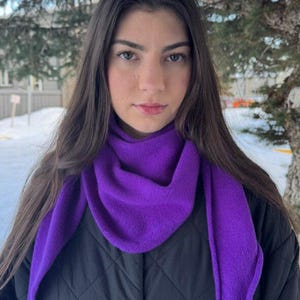 以下が含まれることがあります： 黒いキルティングジャケットを着た女性が、鮮やかな紫色のスカーフを着用しています。 スカーフは首と肩にかけられ、三角形の形をしています。 背景には、木々のある雪景色が広がっています。