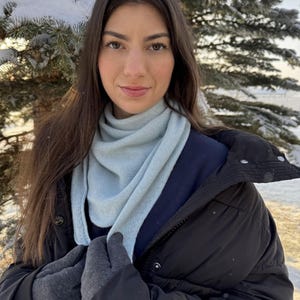 以下が含まれることがあります： 水色のスカーフと黒い手袋を着用した女性、黒いジャケットを着ています。スカーフは首と肩にかけられています。背景には雪の降る常緑樹があります。