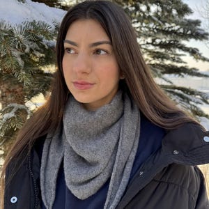 May include: A grey scarf draped around the neck. The scarf is made of a soft material and has a triangular shape. The person is wearing a black jacket and a navy blue sweater. The background includes snow-covered evergreen trees.