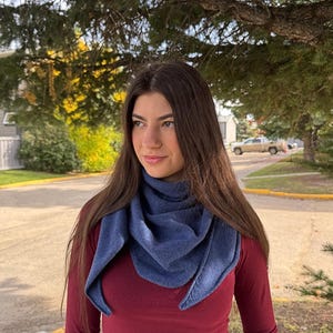 May include: A solid blue triangular scarf with a textured knit fabric draped around the neck, worn over a long-sleeved maroon shirt. The scarf features pointed ends that hang down the chest, creating a layered appearance with soft folds.