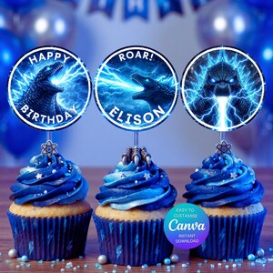 May include: Three cupcakes with blue frosting and Godzilla-themed toppers. The toppers feature Godzilla illustrations with lightning effects and text that reads "Happy Birthday" and "Roar! Elison." The cupcakes are decorated with silver sprinkles.