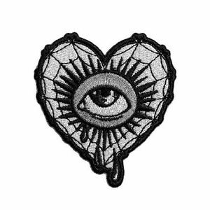 May include: Black and white embroidered heart-shaped patch. The design includes an eye at the centre, encircled by sun rays and a spiderweb pattern. A drop of liquid is shown dripping from the bottom of the heart.
