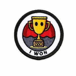 Puede incluir: Un parche circular con borde negro e interior blanco. Presenta un trofeo dorado con una cara sonriente, sobre un fondo rojo y gris. El texto "I WON" está impreso en blanco.