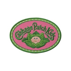 May include: An oval embroidered patch featuring a pink background with a green border containing white dots. The text Cabbage Patch Kids is stitched in green above a central illustration of a baby face nestled within green cabbage leaves.