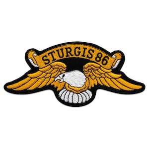 Puede incluir: Un parche bordado que muestra un águila de cabeza blanca con alas doradas extendidas. Una cinta dorada curva se sitúa sobre el águila, mostrando el texto negro STURGIS 86. Todo el diseño está delineado con hilo negro sobre fondo negro.
