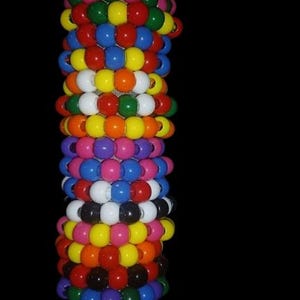 May include: A cylindrical stack of colorful beaded bracelets. The bracelets are made of round beads in various colors, including pink, green, yellow, red, blue, orange, white, and black. The bracelets are stacked on top of each other.