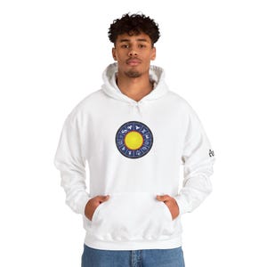 Könnte beinhalten: Weißer Kapuzenpullover mit einem kreisförmigen Design mit einer gelben Sonne und Tierkreiszeichen. Das Design ist auf der Brust zentriert. Der Pullover hat eine Vordertasche und eine Kapuze mit Kordelzug. Die Ärmel sind lang und die Bündchen gerippt.