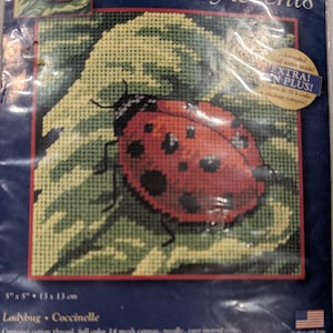 May include: A needlepoint kit by Creative Accents featuring a ladybug on a leaf. The kit includes color-coded thread, a 5" x 5" (13 x 13 cm) canvas, needle, and instructions. The ladybug is red with black spots, and the leaf is green.