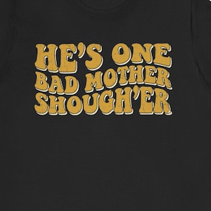 Może przedstawiać: Czarny t-shirt z napisem "HE'S ONE BAD MOTHER SHOUGH'ER" w stylu retro, pomarańczową i żółtą czcionką. Koszulka ma okrągły dekolt i wydaje się być wykonana z miękkiego materiału. Tekst jest wyśrodkowany na przodzie koszulki.
