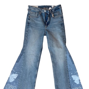 Op de afbeelding: Lichtblauwe denim jeans met een uitlopend beenontwerp. De jeans hebben een vlinder- en hemels ontwerp op de uitlopende panelen. De jeans hebben een knoop- en ritssluiting, met een klassieke vijf-zakken-stijl.