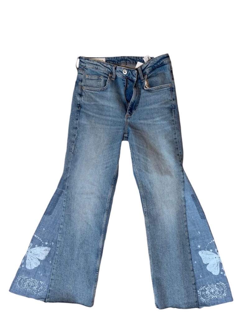Op de afbeelding: Lichtblauwe denim jeans met een uitlopend beenontwerp. De jeans hebben een vlinder- en hemels ontwerp op de uitlopende panelen. De jeans hebben een knoop- en ritssluiting, met een klassieke vijf-zakken-stijl.