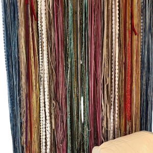 May include: A colorful wall hanging featuring numerous strands of varying textures and colors. The strands include blue, red, yellow, and white, creating a vibrant and textured display. The piece appears to be handmade, adding a unique touch to any space.
