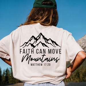 May include: Cream-colored t-shirt with a black mountain graphic and the text "FAITH CAN MOVE Mountains" and "MATTHEW 17:20". The shirt is made of a soft material.