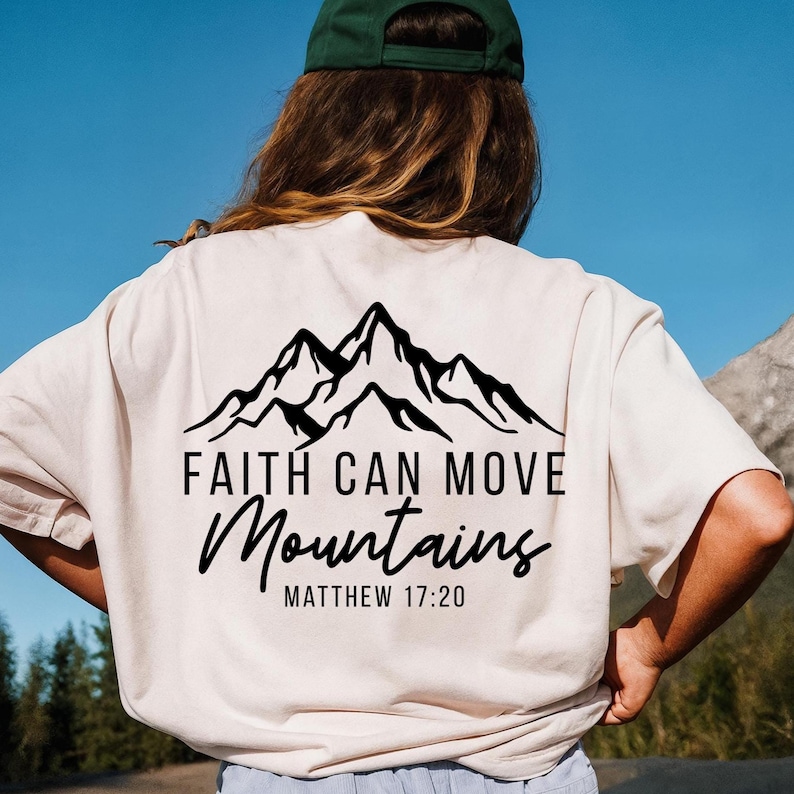 May include: Cream-colored t-shirt with a black mountain graphic and the text "FAITH CAN MOVE Mountains" and "MATTHEW 17:20". The shirt is made of a soft material.