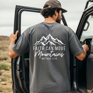 May include: Dark gray t-shirt with a white mountain graphic and the text "Faith Can Move Mountains" and "Matthew 17:20". The shirt is made of a soft material.