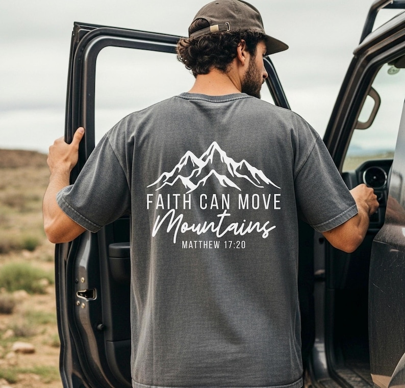 May include: Dark gray t-shirt with a white mountain graphic and the text "Faith Can Move Mountains" and "Matthew 17:20". The shirt is made of a soft material.