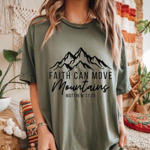 May include: Olive green t-shirt with a black mountain graphic and the text "Faith Can Move Mountains" and "Matthew 17:20". The shirt has short sleeves and a relaxed fit.