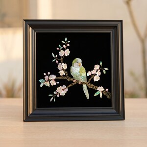 May include: A framed artwork featuring a bird perched on a branch with delicate pink blossoms. The bird has green and white feathers, set against a black background. The frame is black.