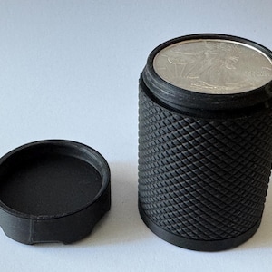 Puede incluir: Un portamonedas negro texturizado con una moneda de plata visible en el interior. El soporte es cilíndrico con un patrón de diamantes y una tapa plana separada. La moneda es un American Eagle de plata.