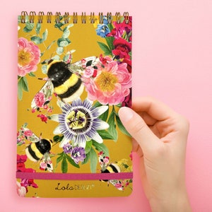 May include: A yellow notepad with a floral and bee design. The notepad has a pink elastic band and says "Lola Design" on the front.