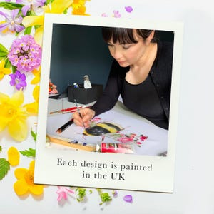 A woman with dark hair paints a bumblebee on a white surface, surrounded by yellow and purple flowers.