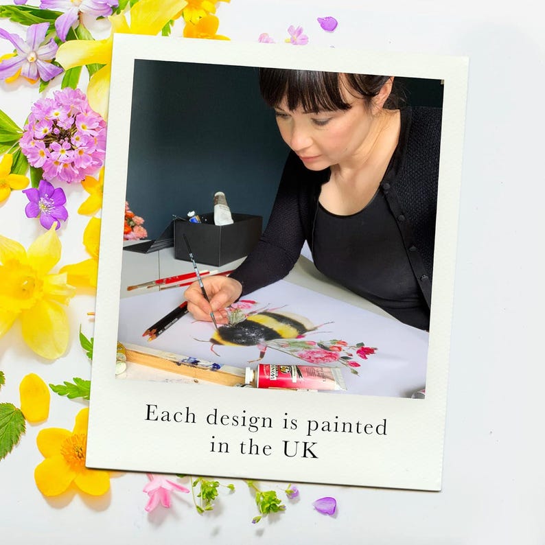 A woman with dark hair paints a bumblebee on a white surface, surrounded by yellow and purple flowers.