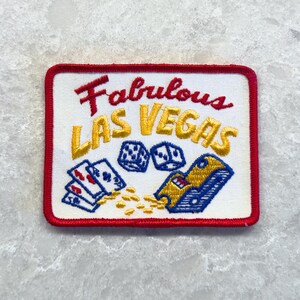 May include: A rectangular embroidered patch with a white background and a red border. It features the red text Fabulous and yellow text LAS VEGAS above illustrations of two blue dice, a hand of playing cards, and a yellow slot machine.