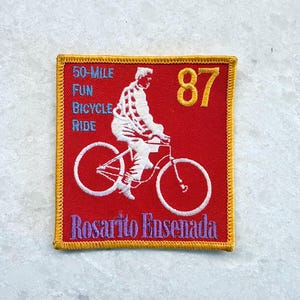 May include: A square embroidered patch with a red background and yellow border. It features a white silhouette of a person riding a bicycle, the number 87 in yellow, and text reading 50-MILE FUN BICYCLE RIDE in blue and Rosarito Ensenada in purple.