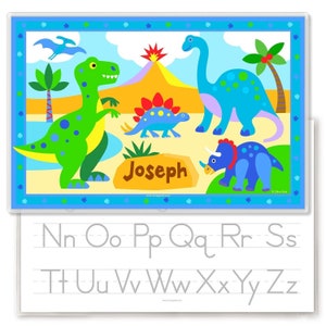 Personalized place mat with fun dinosaur illustrations. Four dinos, palm trees and a volcano in a prehistoric scene. Blue border has dino footprints. Child name in on a rock at the bottom. Reverse side has alphabet letters for tracing.