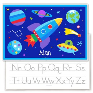 May include: A blue and white dry erase board with a space theme. The board features a rocket, planets, stars, and a UFO. The board also has a space for a name, which is "Alan" in this image. The bottom of the board has a dry erase surface with the letters of the alphabet written in gray.