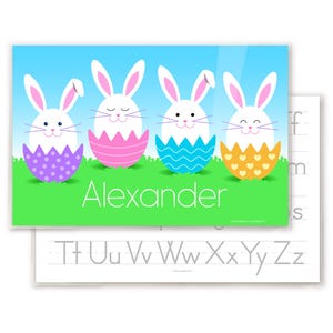 Easter Bunnies place mat for kids with 4 little bunnies, each sitting in a colorful Easter egg shell. Bunnies and eggs are on green grass with blue sky. Child's name is in white letters in grass.